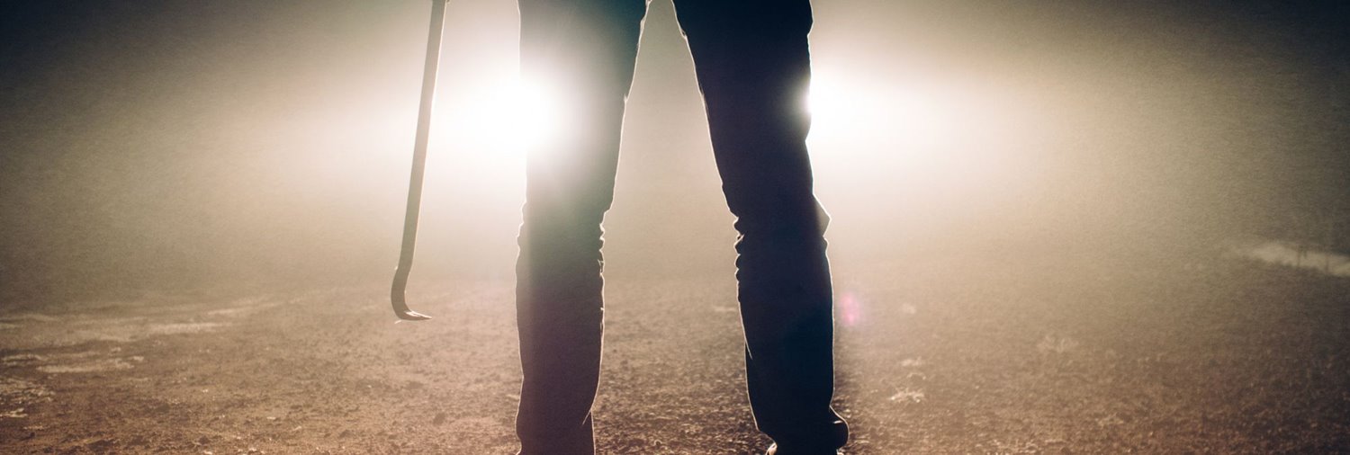 the bottom half of a person holding a crowbar in dark pants backlist by headlights