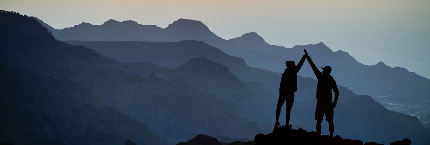 silhouette of two people high-fiving at the top of a peak in the mountains
